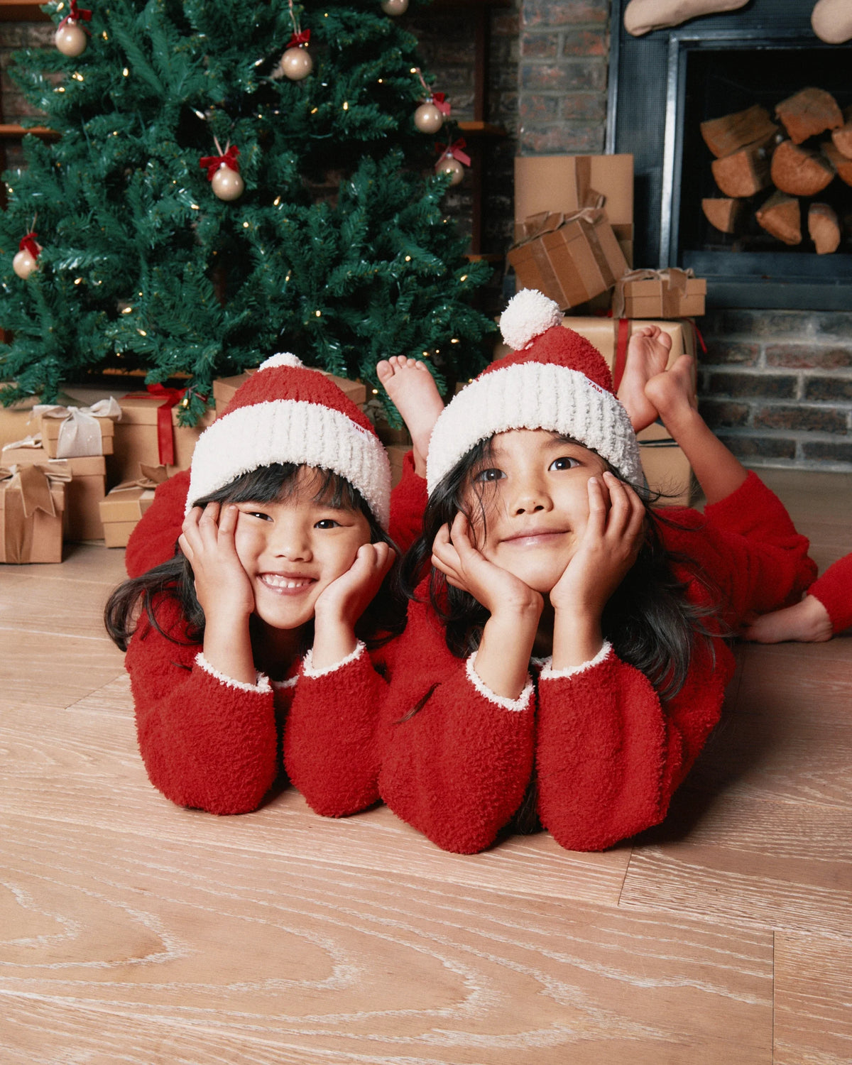 Two little girls laying on the floor and with a christmas tree in the back. They are smiling wearing red beanie, red sweater, red lounge pants