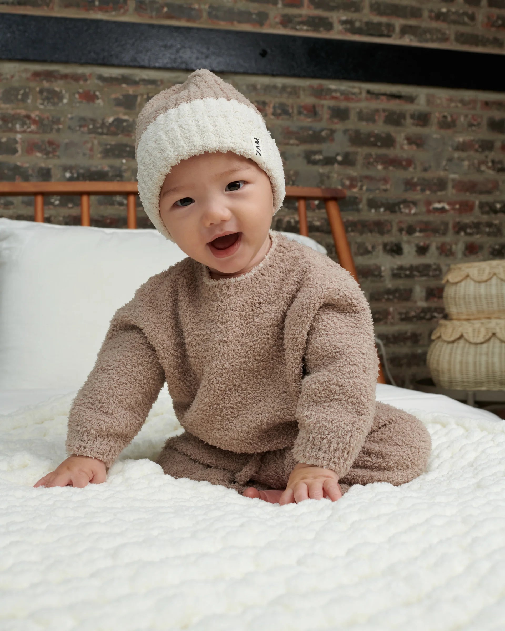 Toddler sitting on a white bed smiling and wearing a Half white half taupe beanie. He is wearing a fuzzy taupe sweater and matching pants.