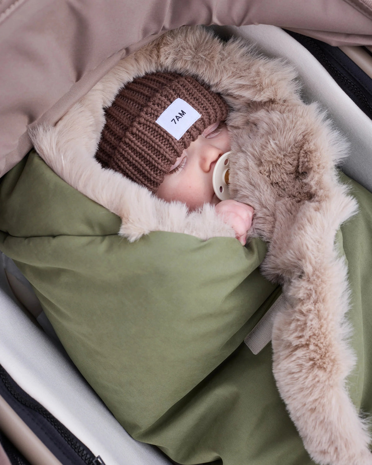 baby sleeping wrapped up in a green blanket with fur inside, wearing a brown beanie that says 7AM.