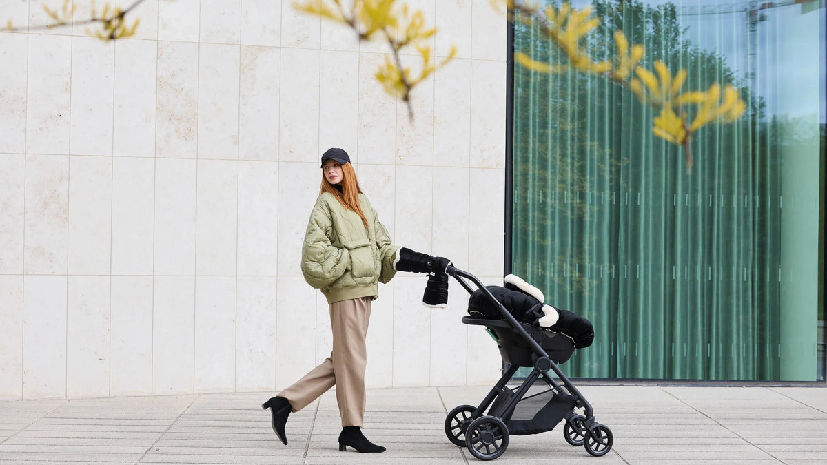 A woman in a light green puffer jacket and beige pants pushes a black stroller along a paved walkway in front of a modern building with a large glass window.