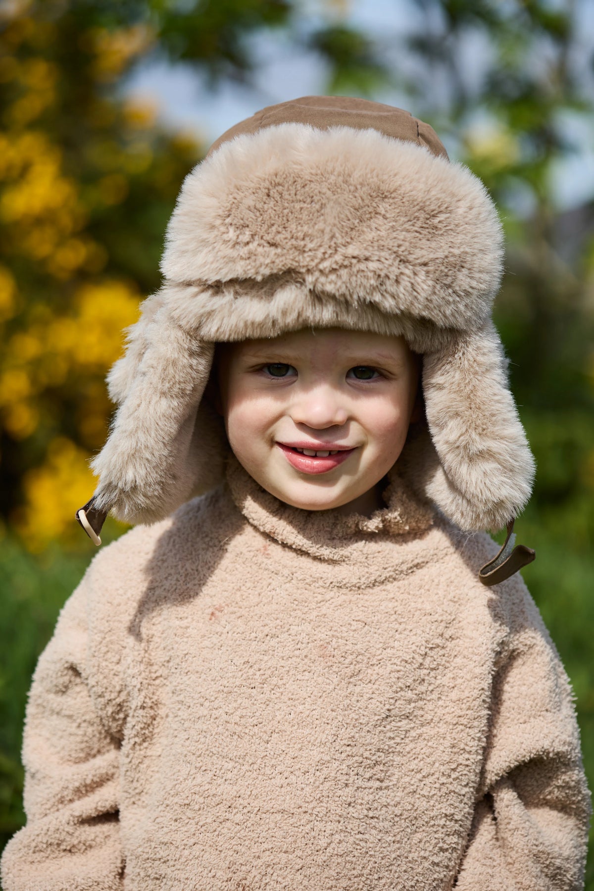 A young child wearing a beige sherpa fleece winter jacket and a matching fluffy fur-trimmed aviator-style hat, photographed outdoors with a blurred green and yellow background.