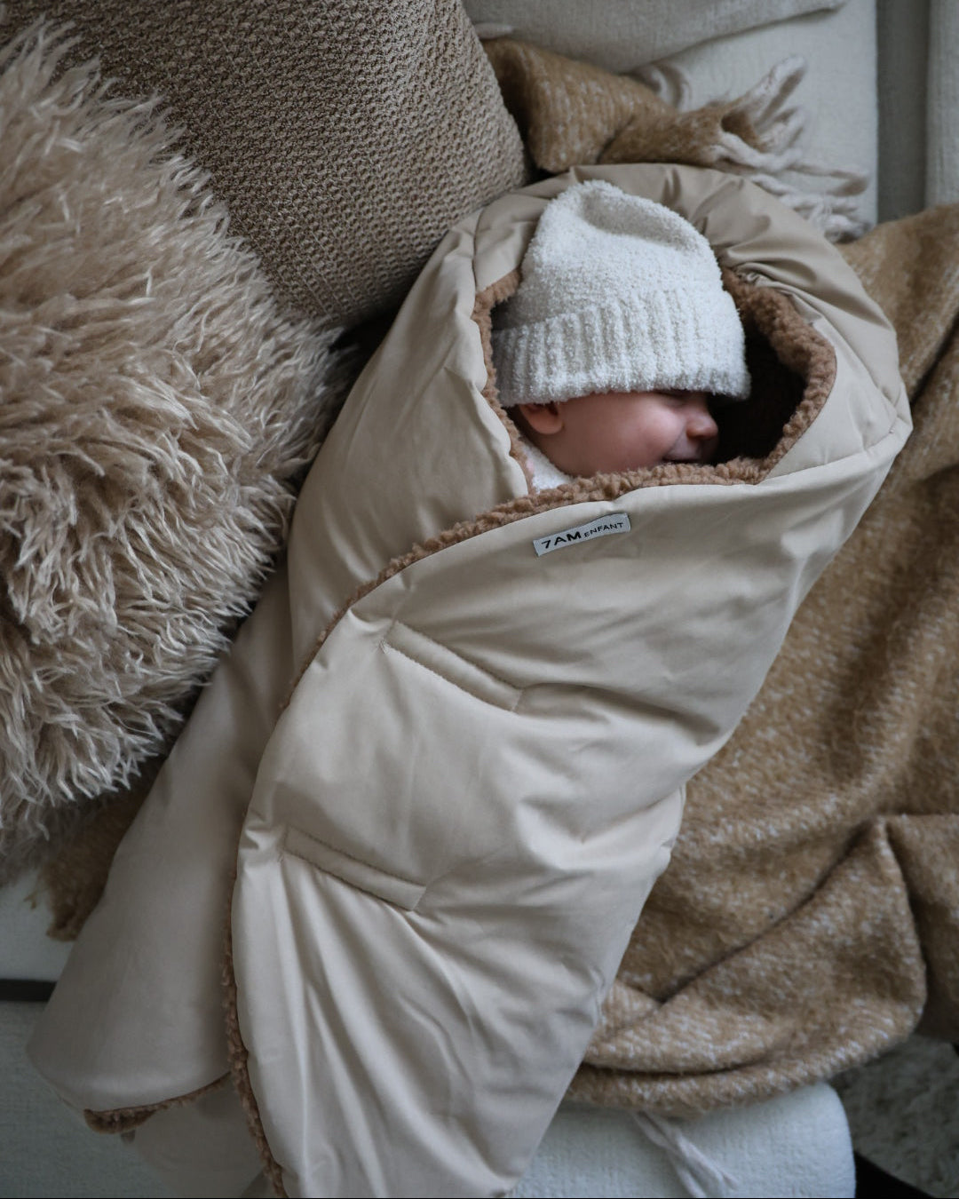 Baby wrapped in a beige swaddle with a white knit hat, sitting on a textured surface.