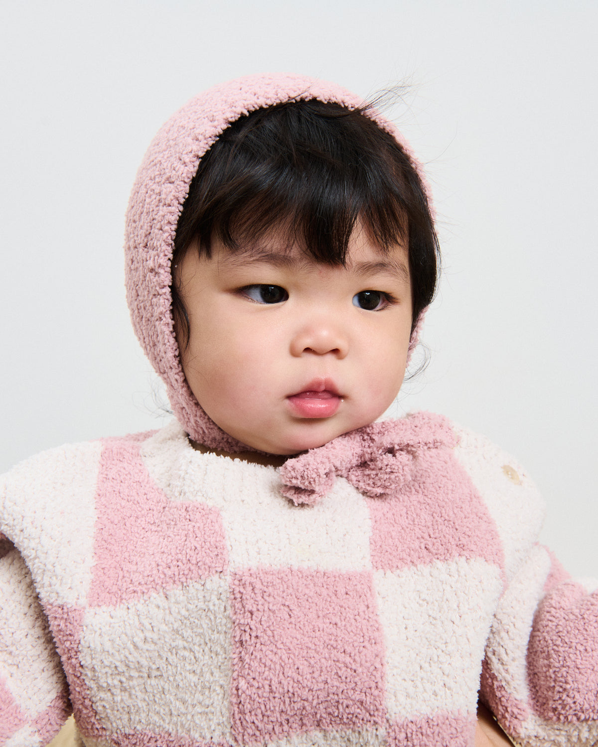 Baby girl wearing a checkered white and pink fuzzy sweater pink bonnet.