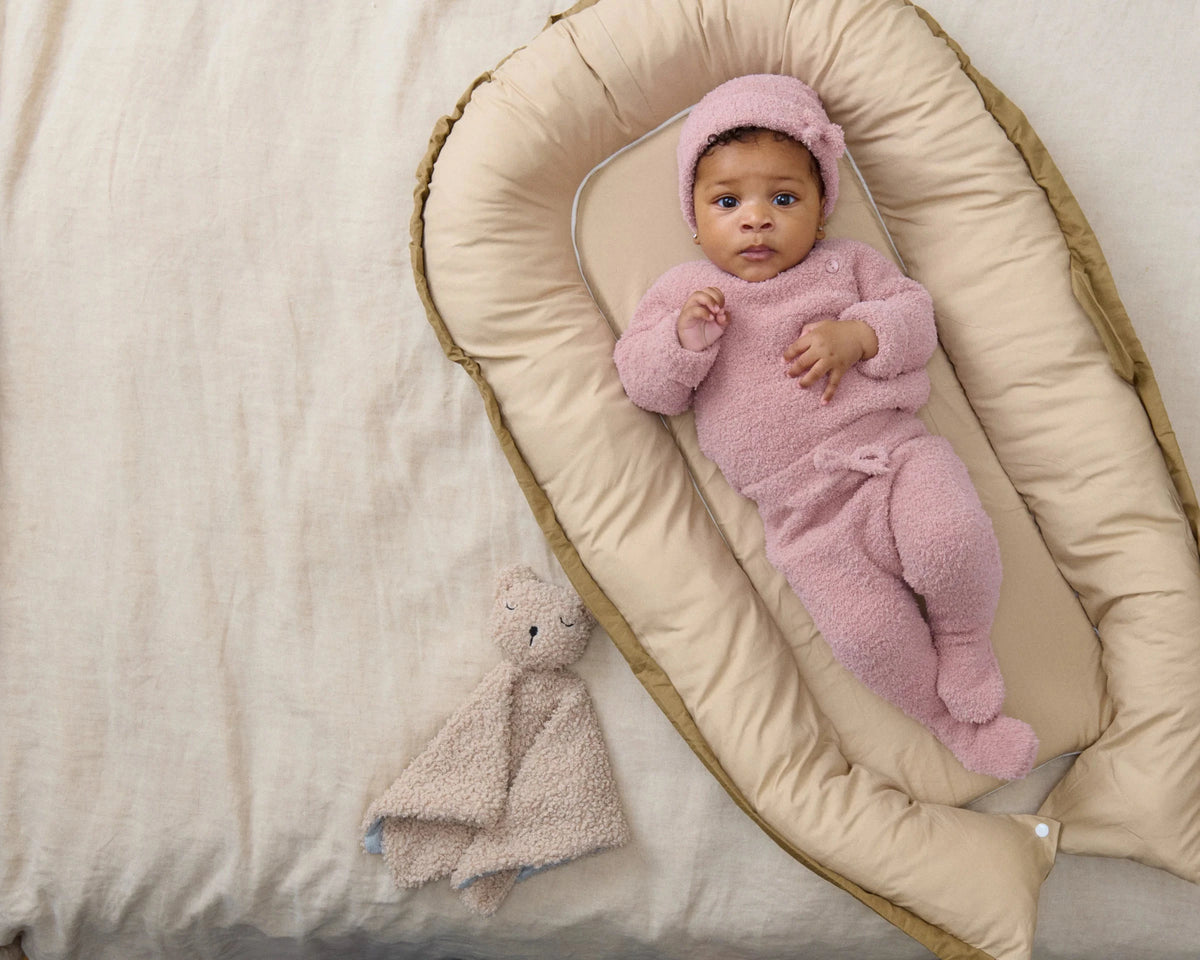 Baby in a pink outfit lying on a beige oval cushion with a soft toy beside.
