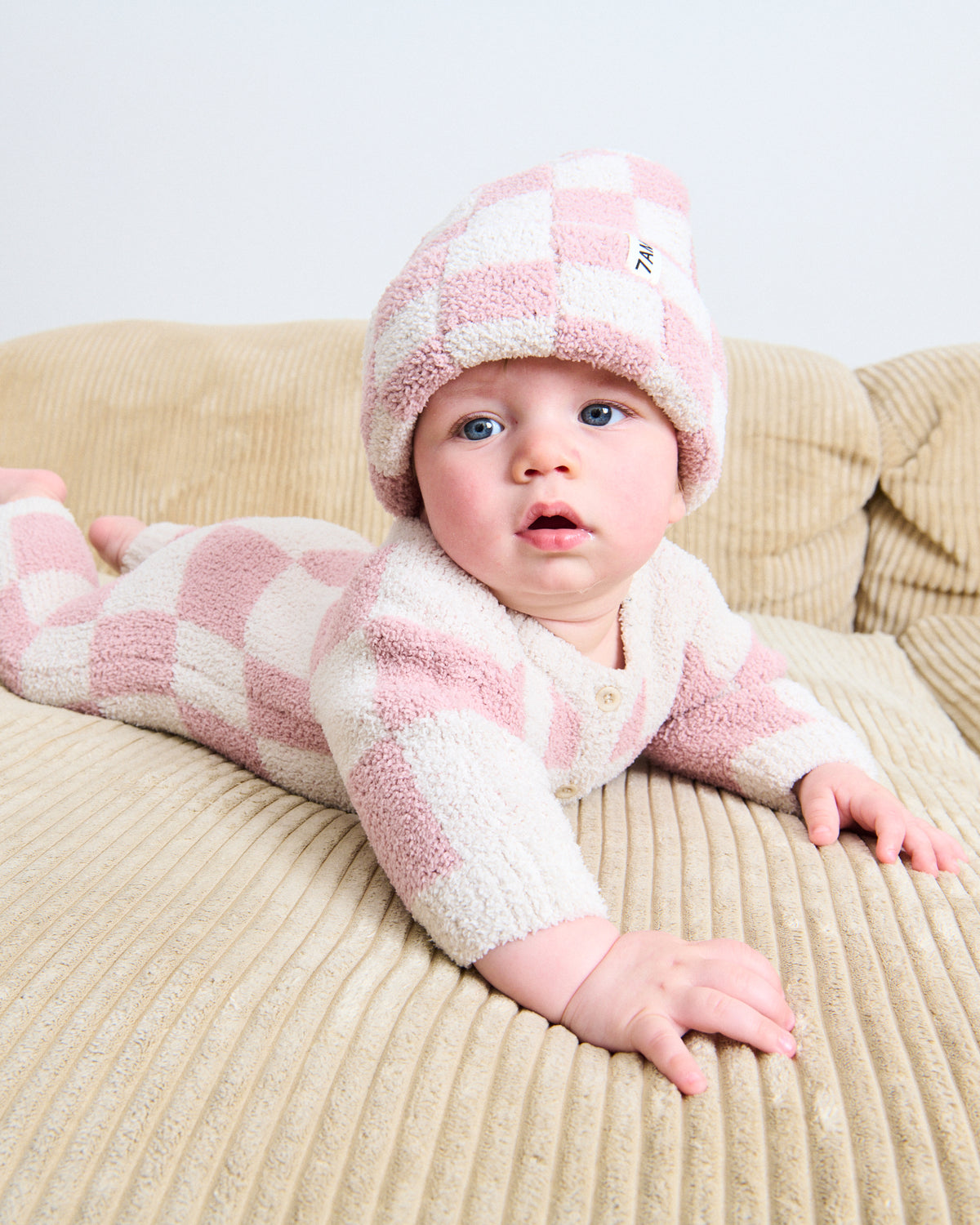 Young baby layed on a couch, wearing a fuzzy checkered beanie in ash rose and ecru, and a fuzzy checkered jumpsuit in ash rose and ecru.