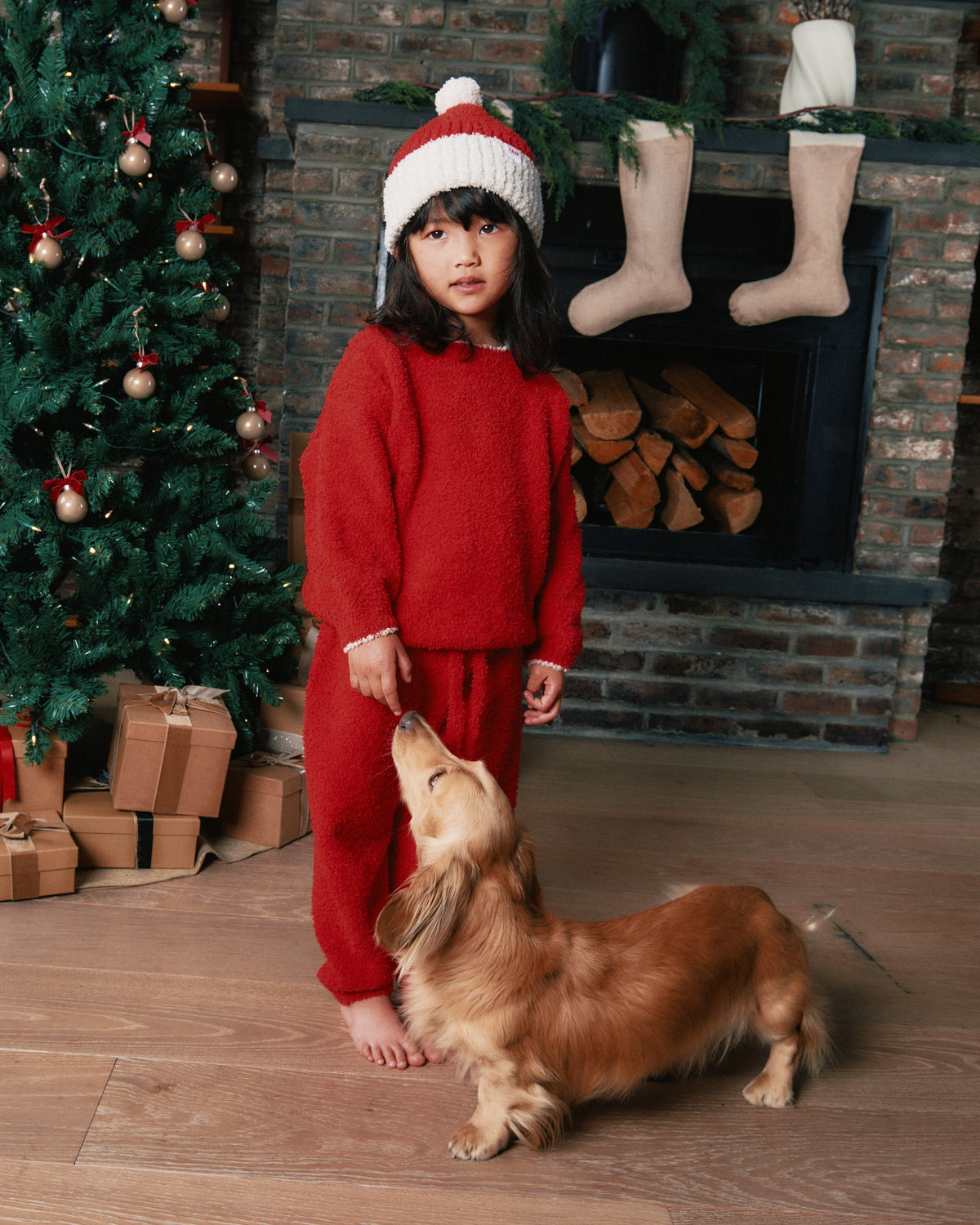 A young child wearing a cozy red knit sweatsuit and a red-and-white Santa hat stands barefoot in a holiday living room, looking up at a small brown dachshund. A decorated Christmas tree, fireplace with logs, stockings, and wrapped gifts are visible in the background.