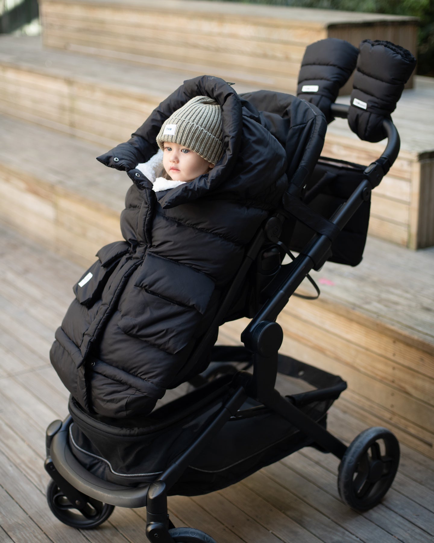 Toddler in a beanie sitting in a puffy black footmuff in a stroller with the background blurred showing wooden steps.