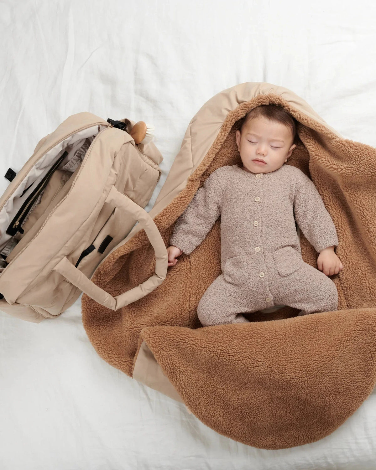 Baby in a brown outfit lying on a beige blanket next to a baby carrier.