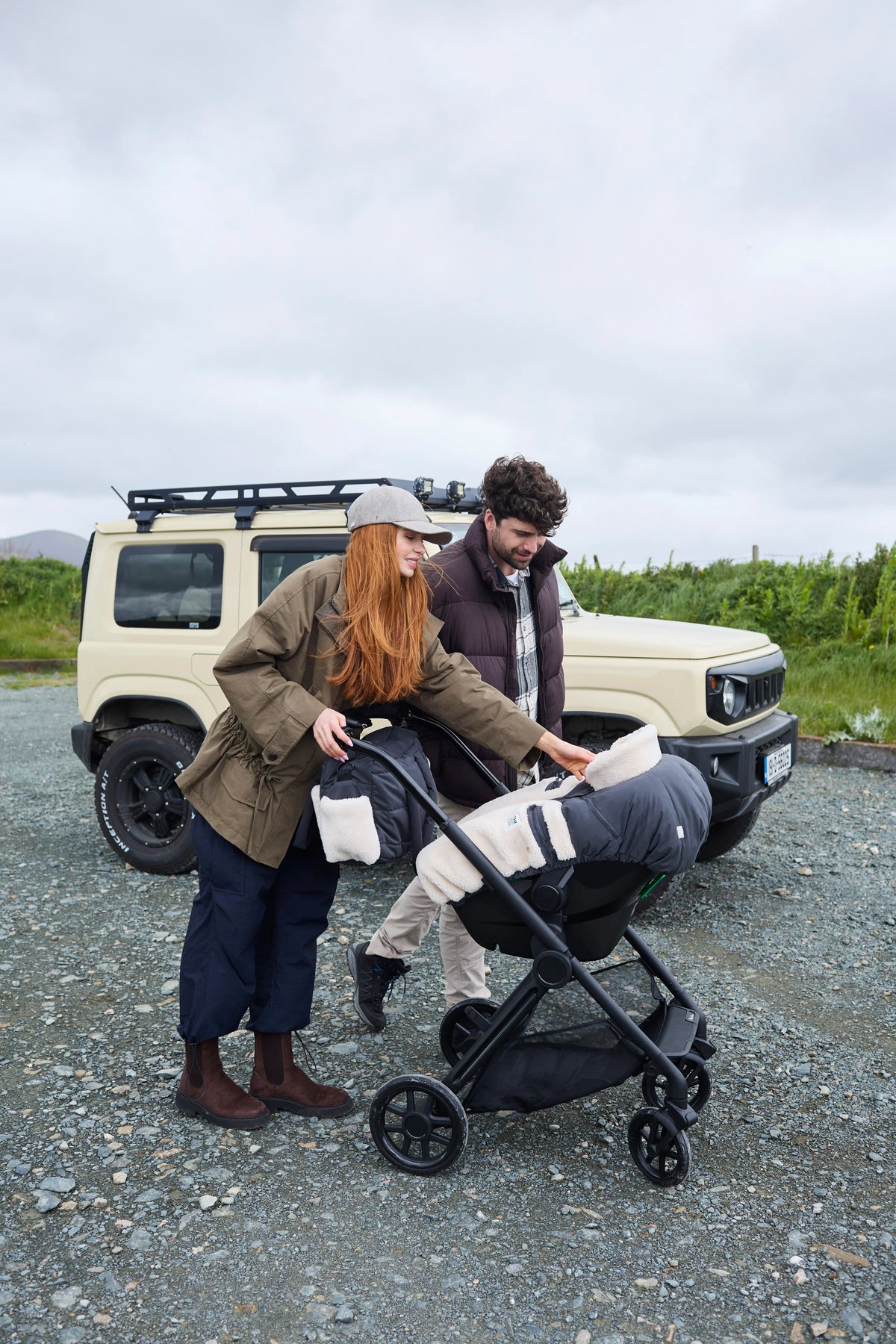 A woman and a man standing outdoors next to a cream-colored SUV, with the woman pushing a black stroller. The woman is wearing a brown jacket, dark pants, and brown boots, and the man is wearing a dark jacket and light pants.