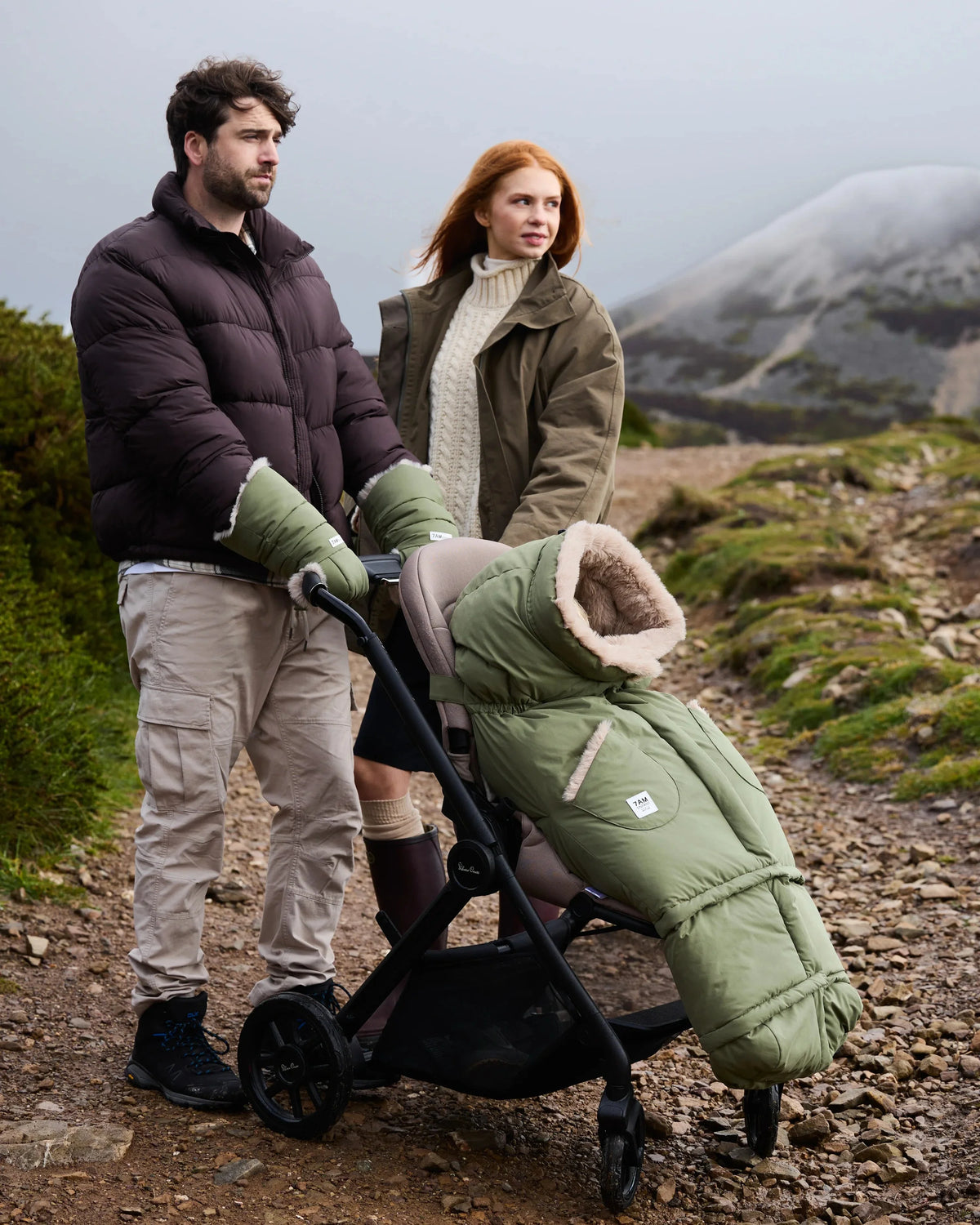 Mom and dad in a mountain background are pushing a stroller with a green foomuff. It is cloudy in the background.