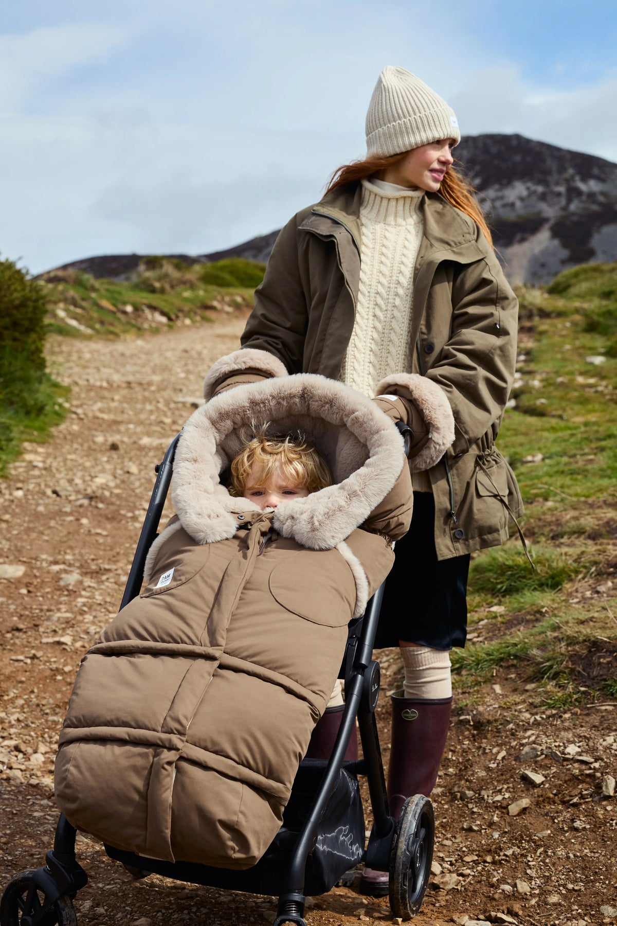 A woman wearing a cream beanie and olive green jacket pushes a stroller along a dirt path outdoors, with a child bundled in a beige quilted puffer cover with faux-fur trim inside the stroller.