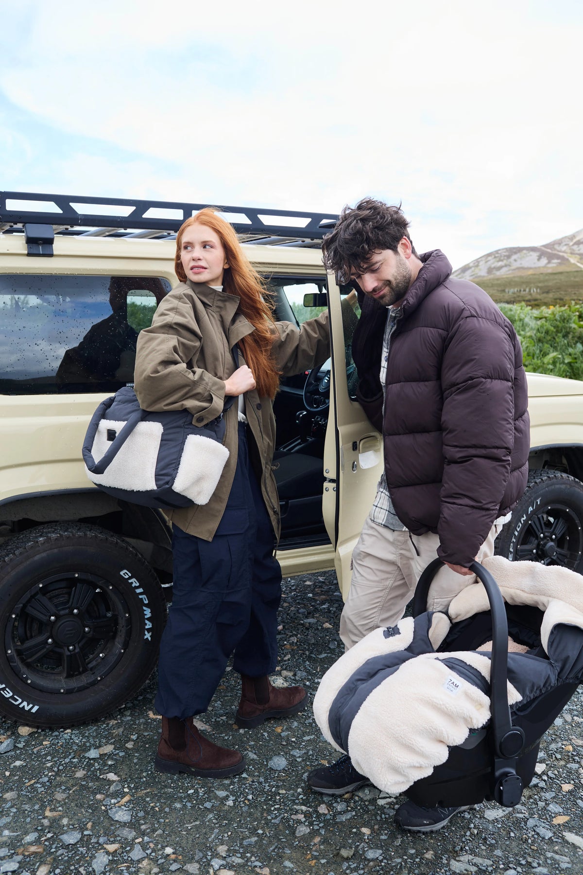 A man and a woman standing outdoors next to a beige off-road vehicle, each carrying a large black and off-white puffer tote bag. The woman wears an olive green jacket and dark pants, and the man wears a dark brown puffer jacket and light pants.