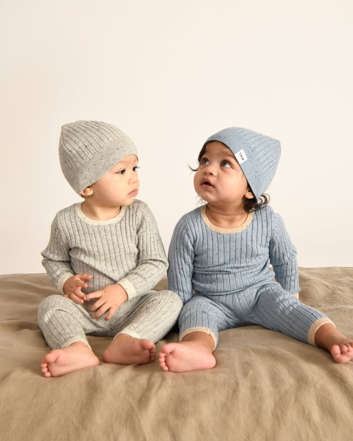 Two babies wearing matching ribbed knit outfits and hats on a beige surface.