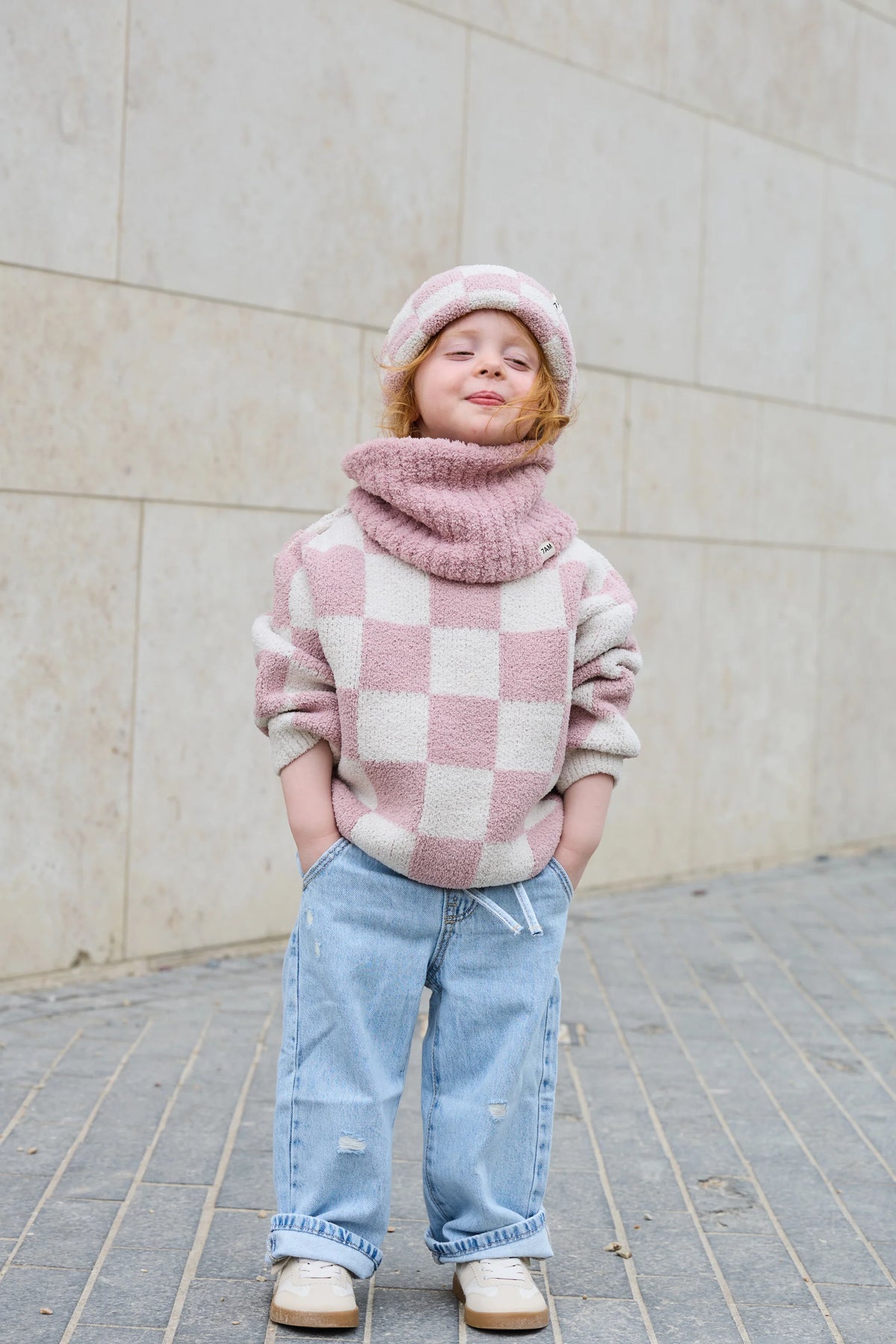 A young child wearing a pink and cream checkered fuzzy sweater, a matching knit beanie, a chunky knit cowl neck warmer, light blue jeans, and beige shoes, standing against a light beige stone wall.