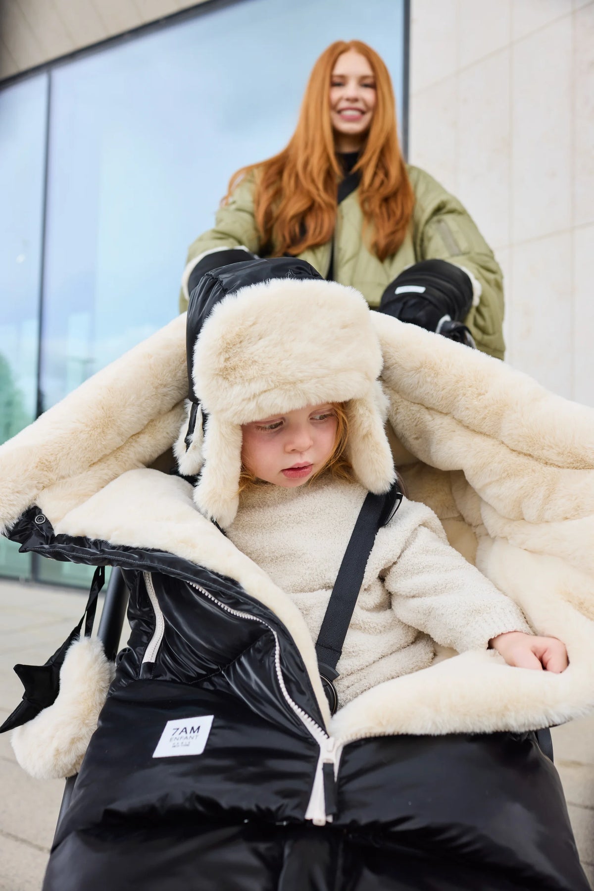 A child wearing a black and cream shearling-lined winter jacket and matching hat, seated in a black puffer stroller, with an adult woman in an olive green coat standing behind them in front of a modern glass building.