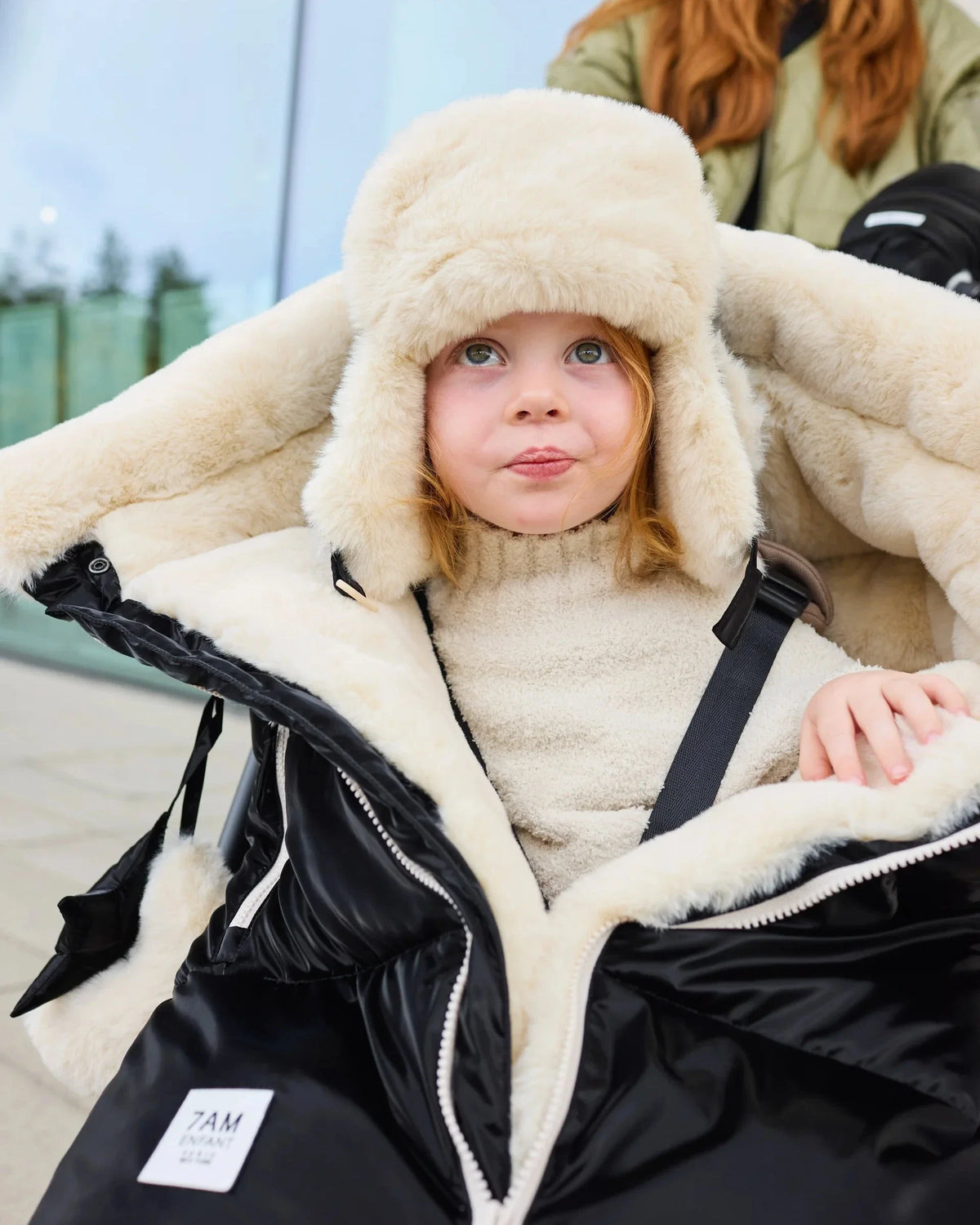 Girl in a stroller wrapped in a white fur footmuff wearing a white fur hat