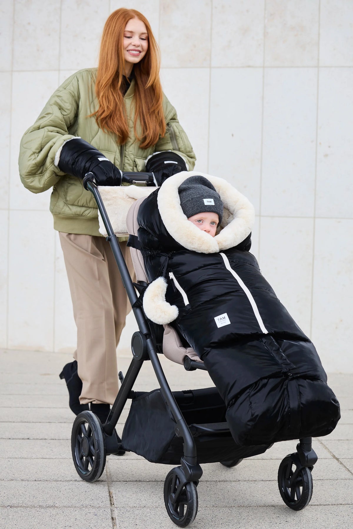 A woman pushes a black stroller outdoors while wearing a black full-body baby winter suit with a cream faux-fur trim. The baby is seated inside the stroller.