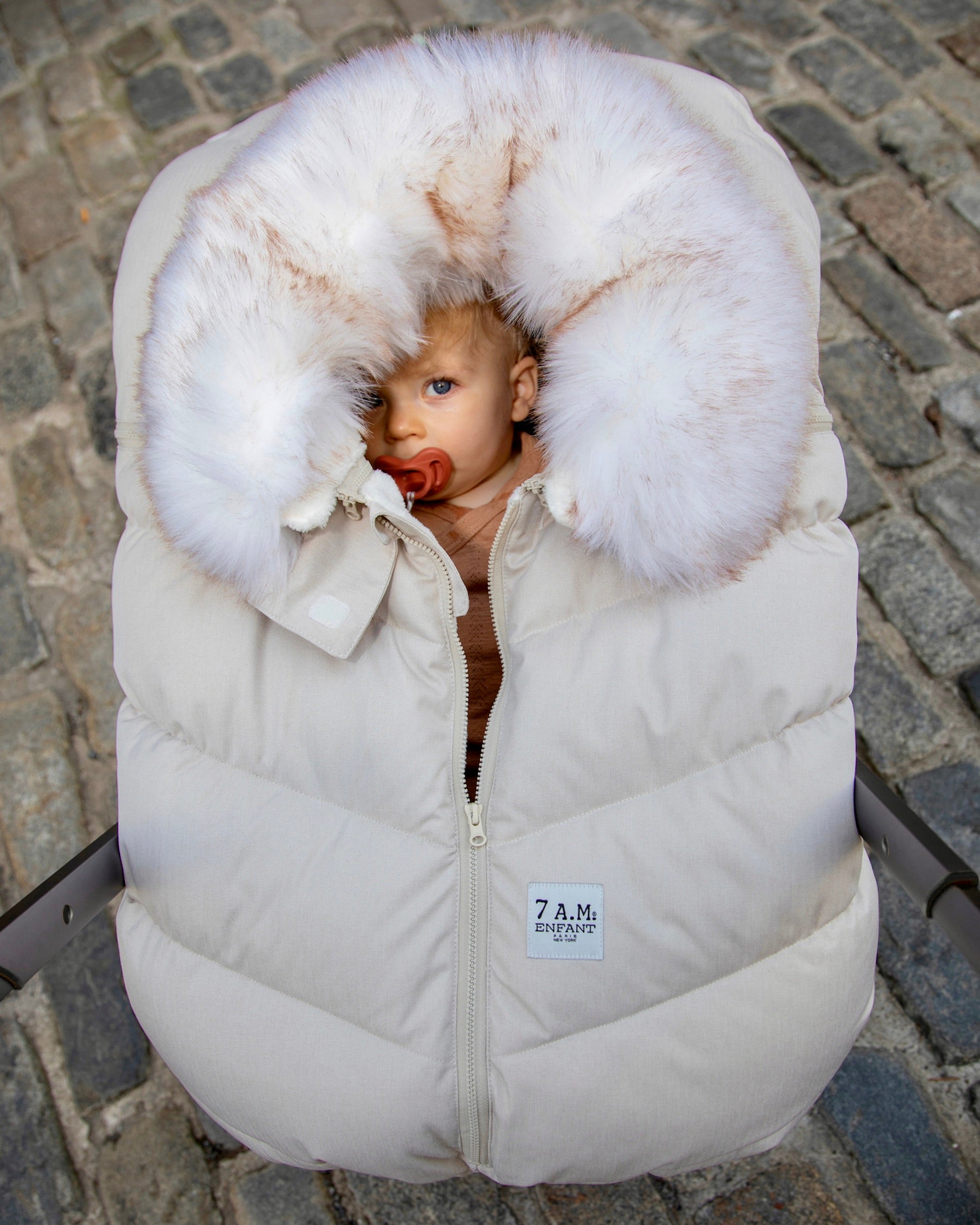 a baby looking to the camera while inside of a stroller, covered by a cream colored stroller cover with a fur collar that surrounds the baby's face. The background is a brick floor.