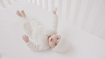 A newborn baby, dressed in a white onesie and hat from the "Fuzzy" collection, is stirring in his crib.